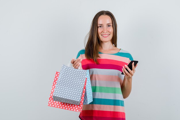 Jonge vrouw met papieren zakken en smartphone in t-shirt en op zoek vrolijk. vooraanzicht.
