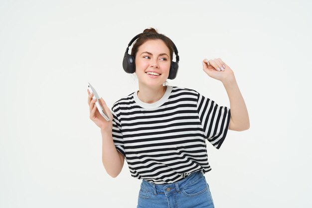 Jonge vrouw met smartphone die naar muziek luistert en danst naar haar favoriete nummer in koptelefoon poseert
