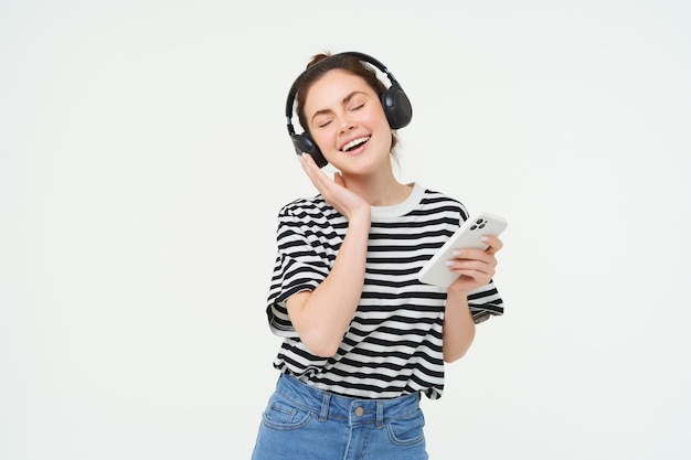 Jonge vrouw met smartphone die naar muziek luistert en danst naar haar favoriete nummer in koptelefoon poseert