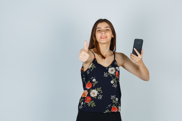 Jonge vrouw met smartphone, duim opdagen in blouse, rok en er vrolijk uitzien. vooraanzicht.