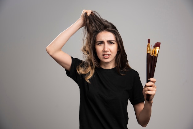 Jonge vrouw met verfborstels wat betreft haar haren op grijze muur.