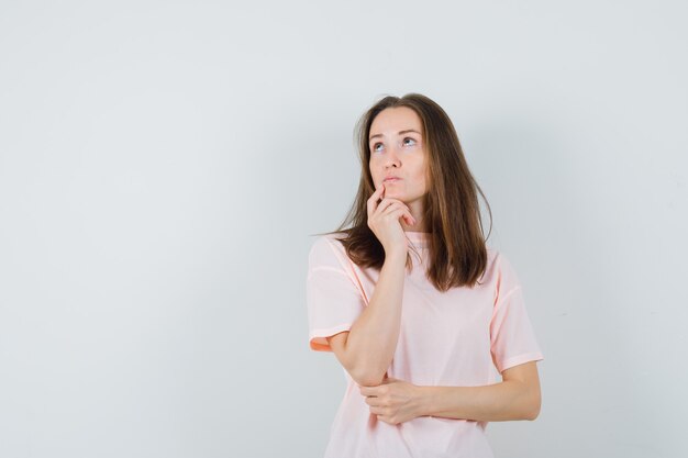 Jonge vrouw met vinger op kin in roze t-shirt en besluiteloos op zoek. vooraanzicht.