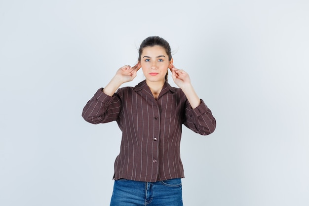 Jonge vrouw met vingers op oren in gestreept shirt, spijkerbroek en serieus, vooraanzicht.