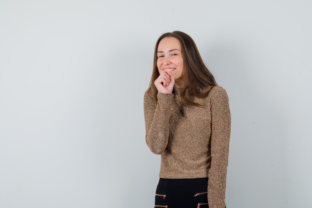 Jonge vrouw met vuist op haar kaak terwijl lachen in gouden blouse en vrolijk kijken