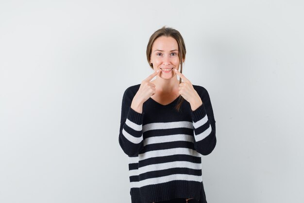 Jonge vrouw met wijsvingers in de buurt van de mond, glimlach in gestreept breigoed en zwarte broek dwingend en gelukkig kijken