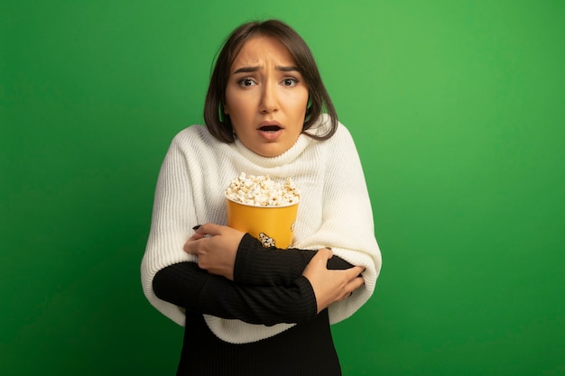 Gratis foto jonge vrouw met witte sjaal met emmer met popcorn bezorgd en verward