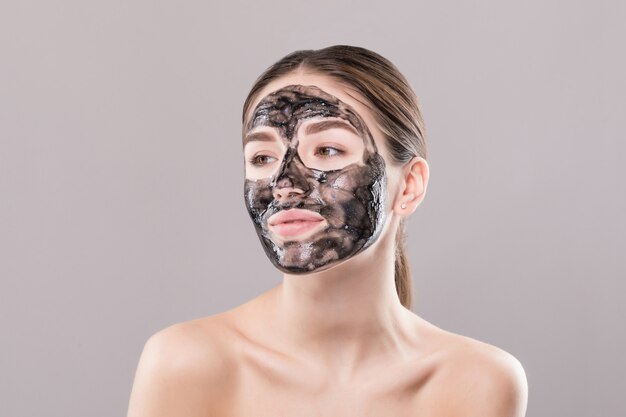 Jonge vrouw met zuiverend zwart masker op haar gezicht dat op witte muur wordt geïsoleerd