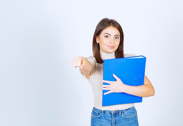 jonge vrouw model houden een blauwe map en wijzend op de camera over witte muur.