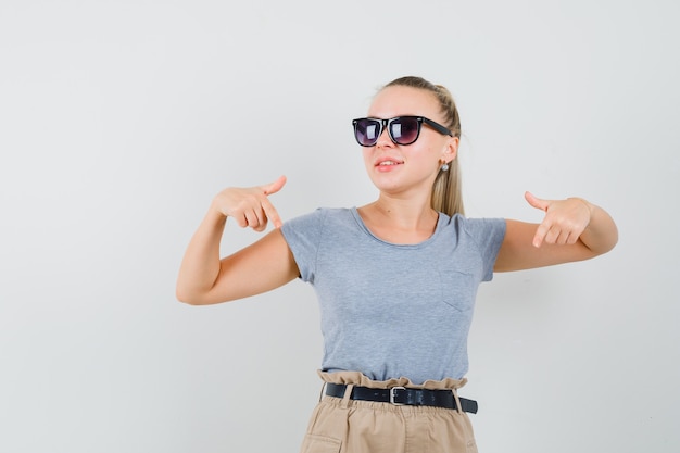 Jonge vrouw naar beneden wijzend in t-shirt, broek en op zoek naar zelfverzekerd, vooraanzicht.