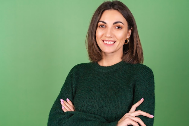 Jonge vrouw op groen in een gezellige grote gebreide trui die schattig lacht met gekruiste handen