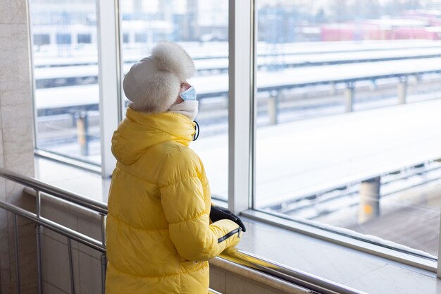 Jonge vrouw op het station in een beschermend masker op het gezicht in winterkleren passagier