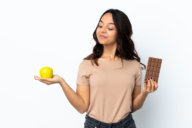 Jonge vrouw over geïsoleerde witte muur die twijfels hebben tijdens het nemen van een chocoladetablet in de ene hand en een appel in de andere