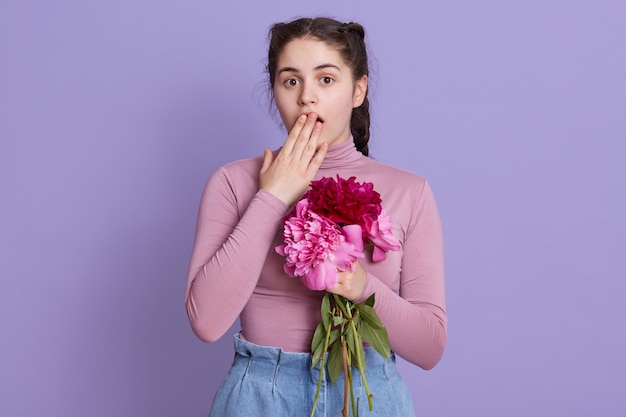 Jonge vrouw over lila muur die casual kleding draagt die bloemen in handen vouwt, geschokt is, mond bedekt met hand voor fout, poseren tegen lila muur.