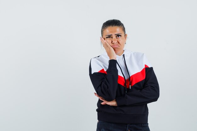 Jonge vrouw permanent in denken pose, grimassen in trui en zwarte spijkerbroek en peinzend, vooraanzicht op zoek.