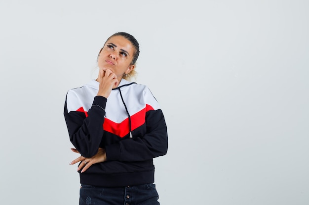 Jonge vrouw permanent in denken pose, hand onder de kin in trui en zwarte spijkerbroek en peinzend op zoek. vooraanzicht.