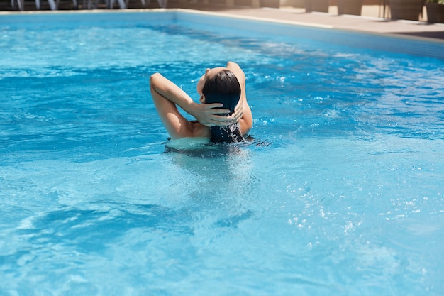 Jonge vrouw poseren achteruit terwijl ze in blauw water staat en haar natte donkere haar aanraakt