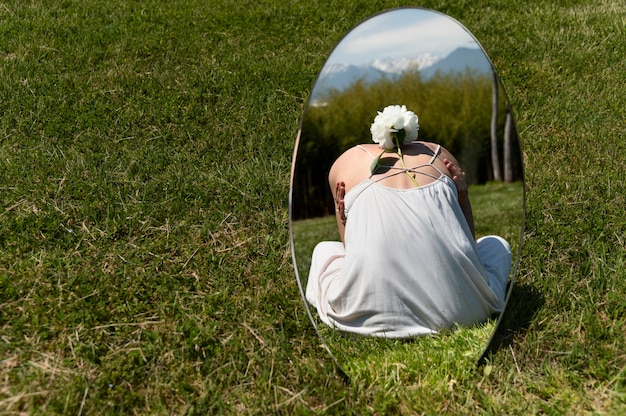 Gratis foto jonge vrouw poseren met pioenroos bloem in de spiegel op het gras buitenshuis