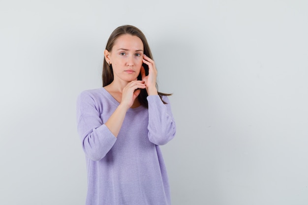 Jonge vrouw poseren terwijl het aanraken van haar gezicht met hand in lila blouse en op zoek verstandig. vooraanzicht.