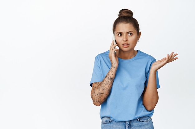 Jonge vrouw praten op mobiele telefoon met verward gezicht, staande in blauw t-shirt op wit