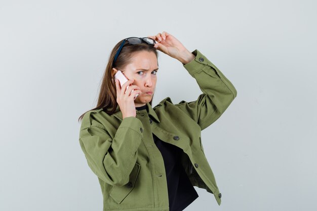 Jonge vrouw praten over de telefoon terwijl het verhogen van de bril boven haar hoofd in groene jas.