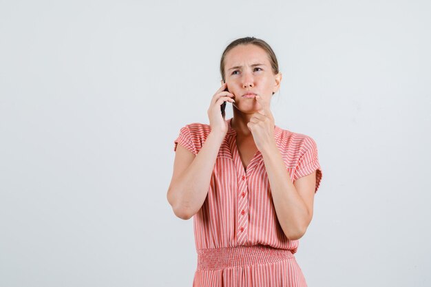 Jonge vrouw praten over mobiel in gestreepte jurk en peinzend, vooraanzicht kijken.