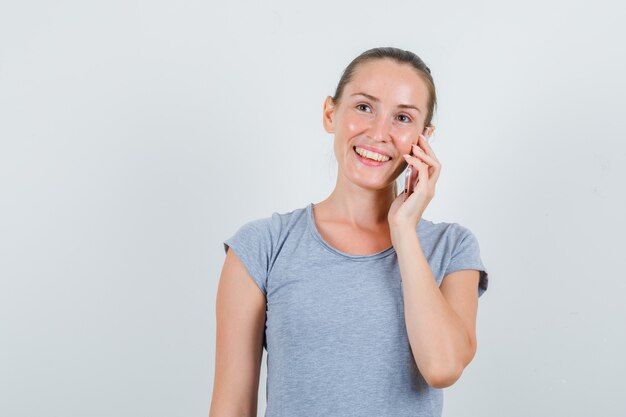 Jonge vrouw praten over mobiel in grijs t-shirt en op zoek blij. vooraanzicht.