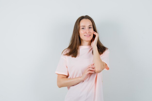 Jonge vrouw raakt haar gezichtshuid op de wang in roze t-shirt aan en kijkt delicaat, vooraanzicht.