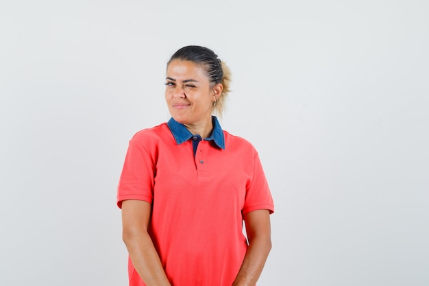 Jonge vrouw rechtop, knipogen en poseren in rood t-shirt en er mooi uitzien. vooraanzicht.