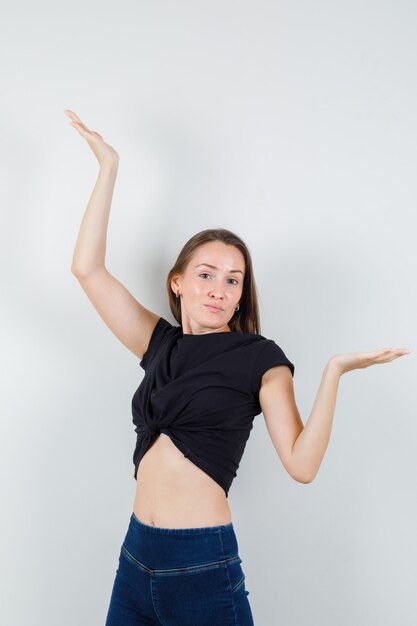 Jonge vrouw schalen gebaar maken in zwarte blouse, broek en op zoek positief.