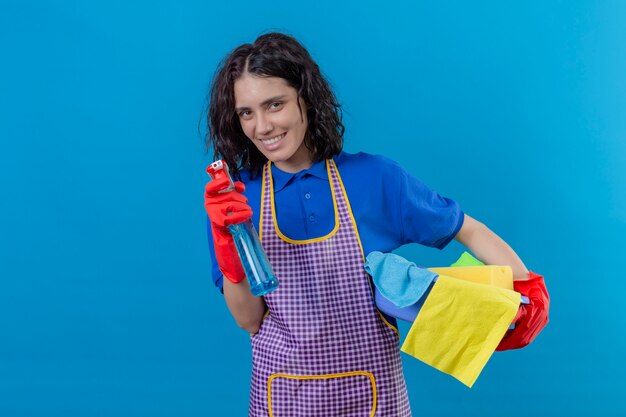 Jonge vrouw schort dragen en rubberhandschoenen die basing met schoonmakende hulpmiddelen houden en nevel schoonmakend die vrolijk over blauwe muur glimlachen