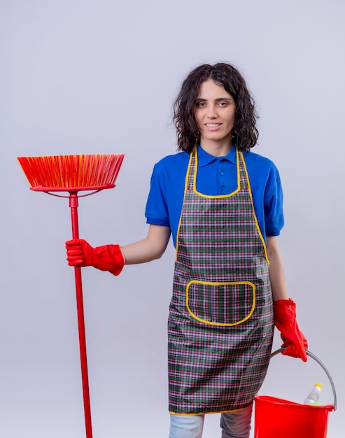 Jonge vrouw schort dragen en rubberhandschoenen die zwabber en emmer met schoonmakende hulpmiddelen met glimlach op gezicht over geïsoleerde witte muur houden