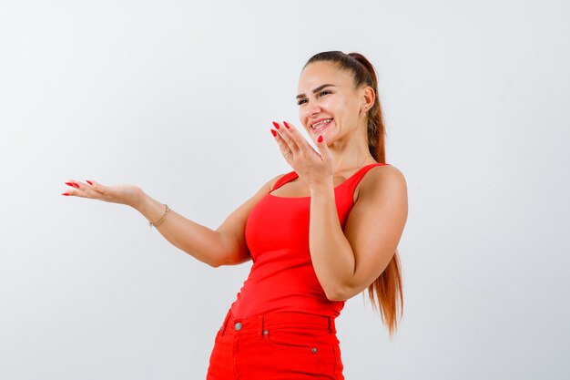 Jonge vrouw spreidt handpalmen in clueless gebaar in rode tanktop, broek en ziet er vrolijk uit. vooraanzicht.