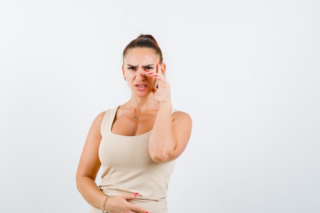 Jonge vrouw staande in denken pose in beige tanktop en besluiteloos op zoek. vooraanzicht.