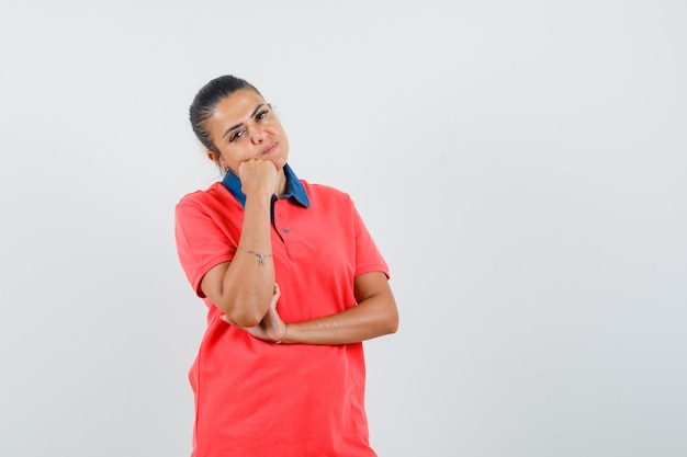 Jonge vrouw staande in denken pose, leunende wang aan kant in rode t-shirt en ziet er mooi uit. vooraanzicht.