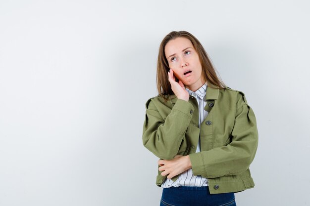 Jonge vrouw staat in denkende pose in shirt, jas en kijkt verveeld. vooraanzicht.