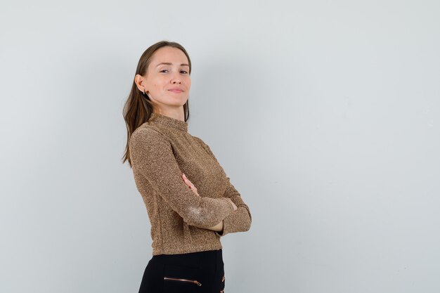 Jonge vrouw staat met gekruiste armen in gouden blouse en kijkt zelfverzekerd, vooraanzicht. ruimte voor tekst