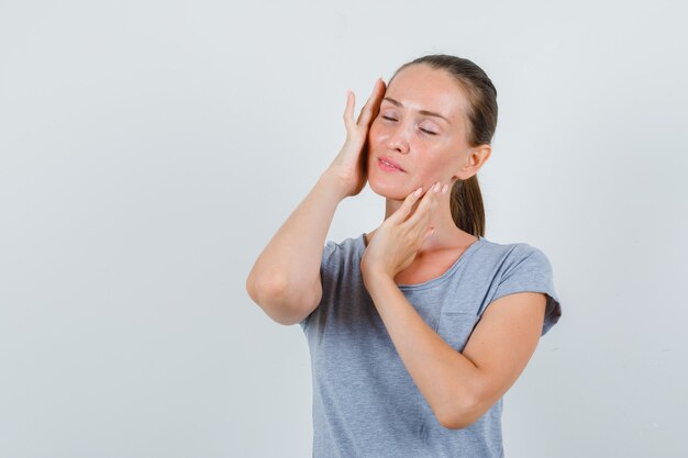 Jonge vrouw tempels met gesloten ogen in grijs t-shirt aan te raken en op zoek vredig, vooraanzicht.