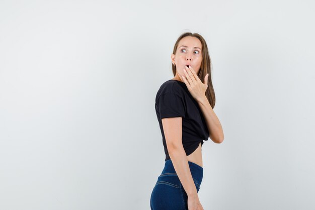 Jonge vrouw terugkijkend met hand op mond in zwarte blouse, broek en verbaasd kijken.