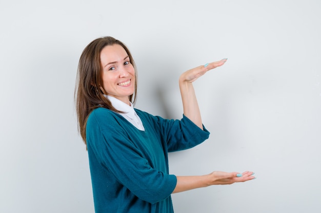 Jonge vrouw toont groot formaat bord in trui over shirt en ziet er vrolijk uit, vooraanzicht.