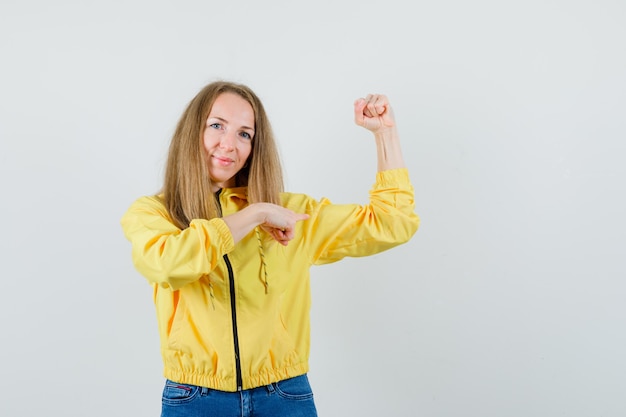 Jonge vrouw toont haar spieren en wijst ernaar in gele bomberjack en blauwe jean en ziet er krachtig uit. vooraanzicht.