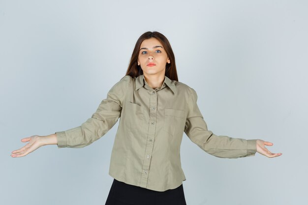 Jonge vrouw toont hulpeloos gebaar door haar schouders op te halen in shirt, rok en verward te kijken. vooraanzicht.