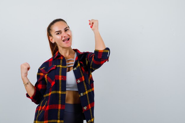 Jonge vrouw toont winnaar gebaar in crop top, geruit hemd en ziet er zalig uit. vooraanzicht.