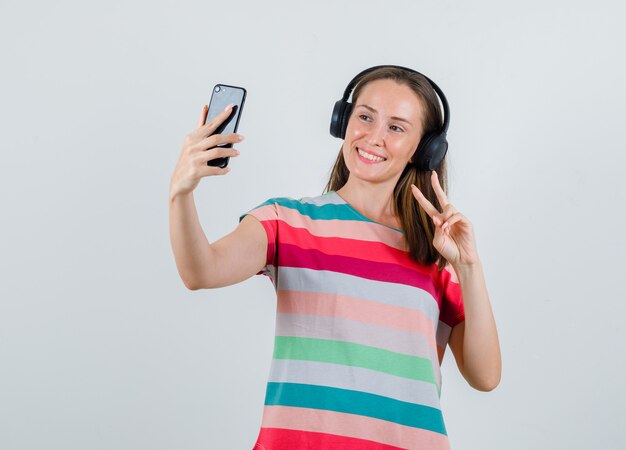 Jonge vrouw v-sign tonen tijdens het nemen van selfie in t-shirt, koptelefoon en op zoek vrolijk, vooraanzicht.