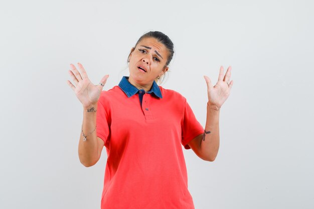 Jonge vrouw verhogen handen in overgave pose in rood t-shirt en op zoek bang, vooraanzicht.