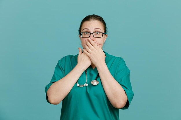 Jonge vrouw verpleegster in medisch uniform met stethoscoop om nek kijken camera geschokt over mond met handen permanent over blauwe achtergrond