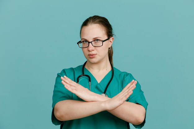 Jonge vrouw verpleegster in medisch uniform met stethoscoop om nek kijken camera met ernstig gezicht maken stop gebaar overschrijding van haar handen permanent over blauwe achtergrond