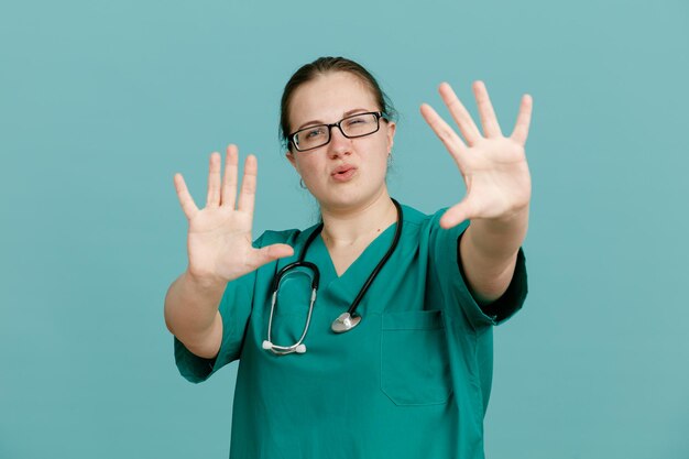 Jonge vrouw verpleegster in medisch uniform met stethoscoop om nek kijken camera met nummer tien met open handpalmen permanent over blauwe achtergrond