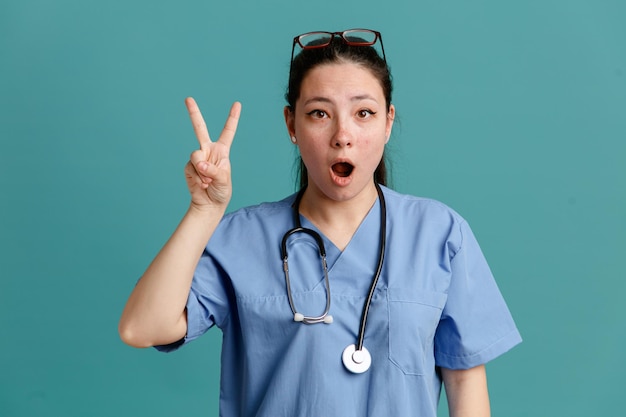 Jonge vrouw verpleegster in medisch uniform met stethoscoop om nek kijken camera verrast met nummer twee met vingers permanent over blauwe achtergrond