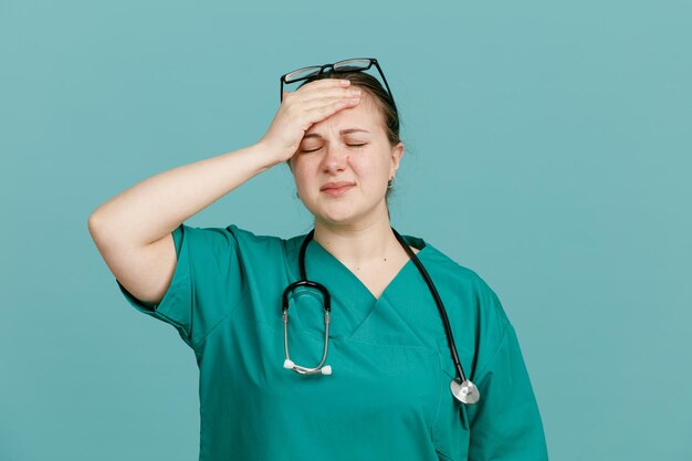 Jonge vrouw verpleegster in medisch uniform met stethoscoop om nek ziet er moe en overwerkt uit met hand op haar voorhoofd staande over blauwe achtergrond