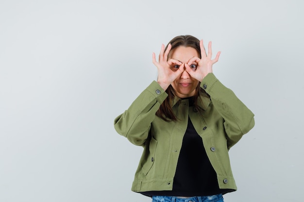 Jonge vrouw verrekijker gebaar in groene jas tonen en op zoek grappig. vooraanzicht.